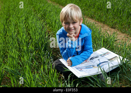 Libre d'un enfant garçon blond lisant un livre à l'extérieur dans un champ de blé, pelouse, jardin, cour, seul, l'étude, l'heure d'été Banque D'Images