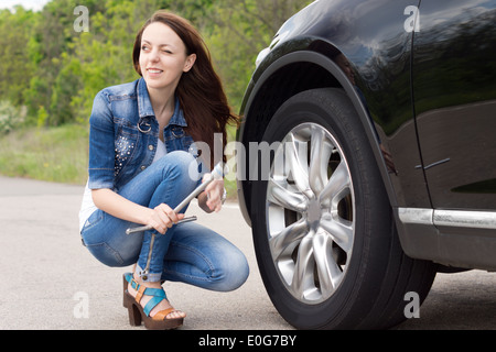 Souriante jeune femme s'apprête à changer un pneu accroupi sur le côté de la voiture avec une clé de roue dans ses mains Banque D'Images