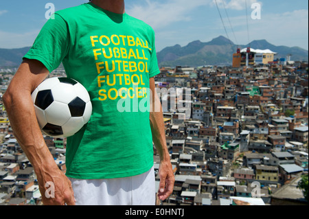 Homme debout dans brésilien de football international t-shirt holding soccer ball en face de taudis favela de Rio de Janeiro d'arrière-plan Banque D'Images