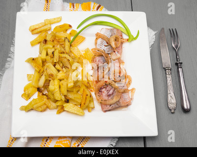 Pommes de terre sautées avec du hareng et l'oignon dans square plaque blanche avec vieux couteau et fourchette sur table en bois gris Banque D'Images