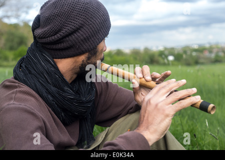 Mixed Race man playing flute dans l'herbe longue sur Hampstead Heath portant des vêtements amples et foulard Banque D'Images