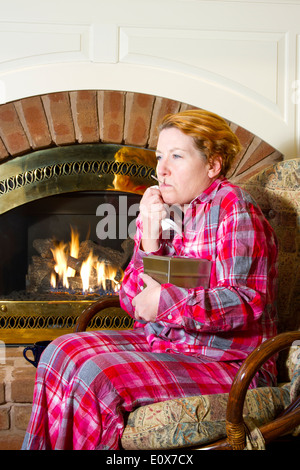 Femme mature habillé en pyjama en flanelle assis sur une chaise en face d'une cheminée dans la maison prendre sa température avec un ou Banque D'Images