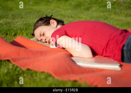 Une belle jeune femme de prendre une sieste relaxante après la lecture d'un livre sur une couverture rouge dans un jardin Banque D'Images