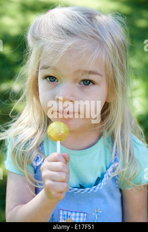 Petit enfant blond girl licking lollipop en extérieur dans jardin, portrait Banque D'Images