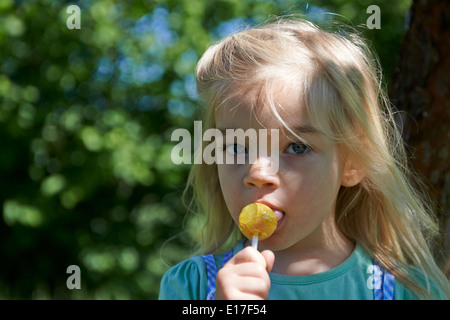 Petit enfant blond girl licking lollipop en extérieur dans jardin, portrait Banque D'Images