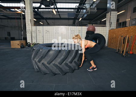 Athlète féminine difficile renversant un énorme pneu. Jeune femme faisant de l'exercice au crossfit gym. Banque D'Images