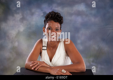 Jolie femme noire en robe blanche, assis avec ses bras sur une table, souriant à la caméra avec plaisir et joie Banque D'Images