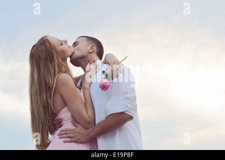 Un jeune couple portrait romantique plage au coucher du soleil s'embrassant. La femelle porte une robe vintage 50s, tenant une rose rose.