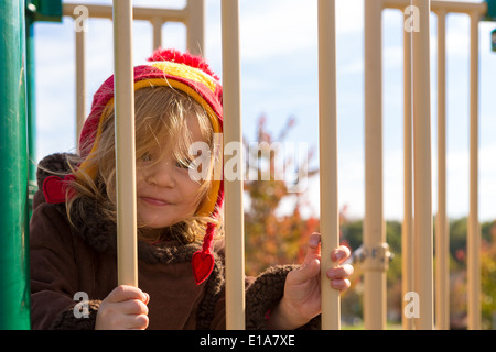 Petite fille à l'aire de jeux vous regarde malicieusement derrière les barres jaunes avec son chapeau d'hiver rouge Banque D'Images