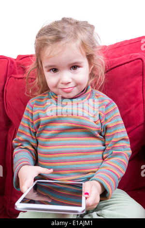 Beau tout-petit avec des cheveux ébouriffés et un adorable sourire assis sur un canapé rouge une tablette ordinateur dans ses mains en sa amuse Banque D'Images