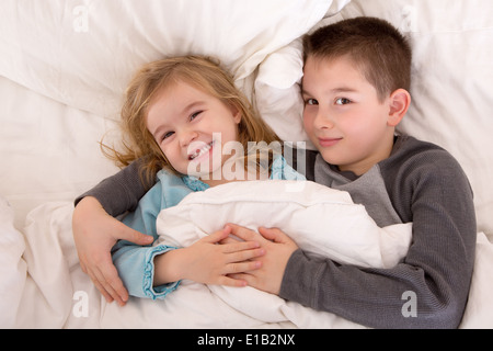 Les jeunes frère et sœur espiègle lit en souriant à la caméra en tant qu'elles se trouvent côte à côte sous la couette sommeil préparation Banque D'Images