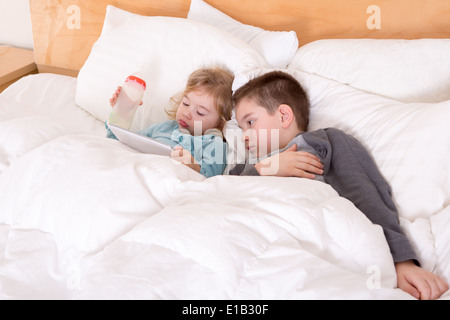 Heureux petit frère et sœur au lit ensemble comme ils câlins sous la couette la lecture d'un ordinateur tablette avant d'aller au s Banque D'Images