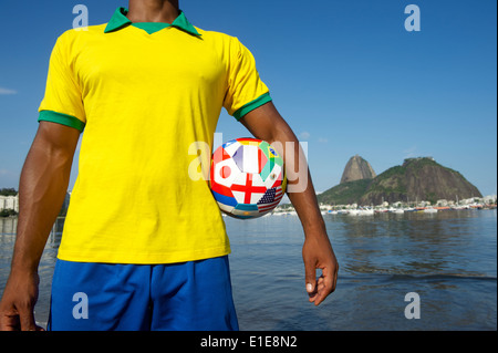 Joueur de football brésilien, comité permanent avec les pays d'un drapeau d'un ballon de football en face de Pao de Acucar Sugarloaf Mountain Banque D'Images
