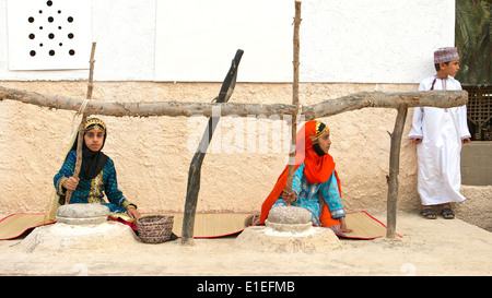 Deux jeunes filles en costume traditionnel omanais le broyage du maïs, tandis que leur frère attend dans l'arrière-plan, à Muscat, Sutanate d'Oman. Banque D'Images