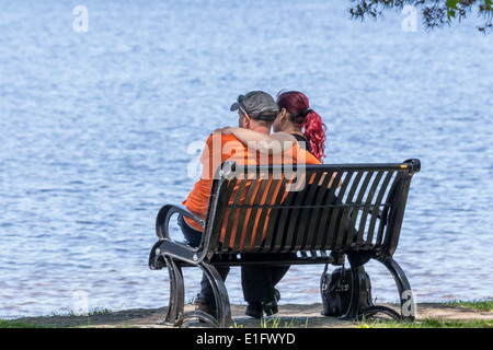 Jeune couple avec les bras autour de l'autre sur le banc de parc donnant sur le lac. Banque D'Images