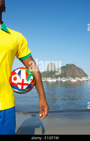 Joueur de football brésilien, comité permanent avec les pays d'un drapeau d'un ballon de football en face de Pao de Acucar Sugarloaf Mountain Banque D'Images