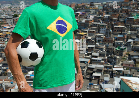 Footballeur Favela standing in Brazilian flag t-shirt holding soccer ball en face de l'arrière-plan des bidonvilles de Rio de Janeiro Banque D'Images