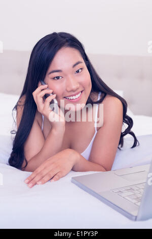Serveuse asiatique smiling at camera lying on bed talking on phone Banque D'Images