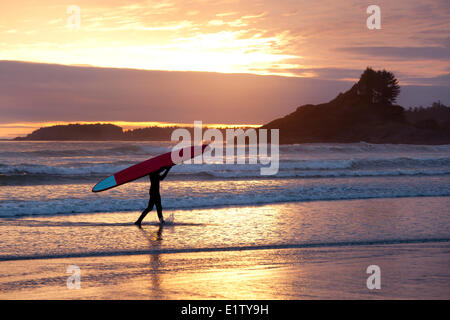 Un internaute porte son surfboard at Cox Bay au coucher du soleil près de Tofino, Colombie-Britannique Canada sur l'île de Vancouver et dans le détroit de Clayoquot Banque D'Images