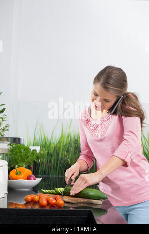 Smiling woman using cell phone tout en réduisant le concombre au comptoir de la cuisine Banque D'Images