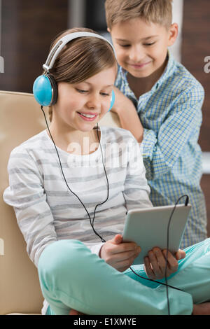 Jolie fille à l'écoute de la musique sur tablette numérique lorsque frère debout derrière elle à la maison Banque D'Images