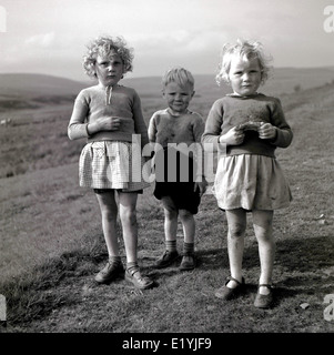 1950 portrait historique par J Allan Paiement de trois jeunes enfants du pays se tenant ensemble sur une colline herbeuse. Banque D'Images