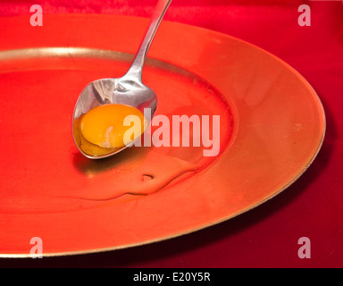 Le jaune d'œuf sur une cuillère en argent reposant sur une plaque rouge cerclée d'or Banque D'Images