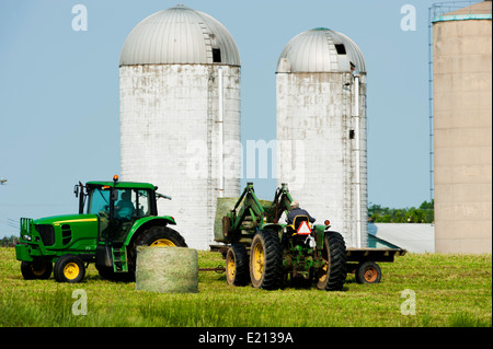 Farmer Chargement des bobines de foin en balles rondes sur un tracteur avec remorque à plateau, avec les fermes silos dans l'arrière-plan Banque D'Images