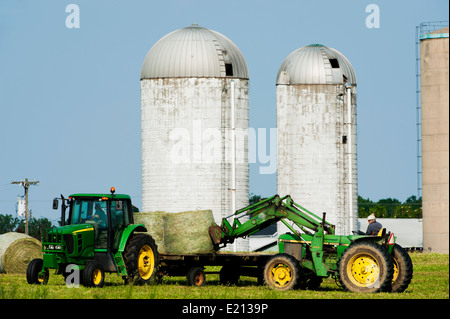 Farmer Chargement des bobines de foin en balles rondes sur un tracteur avec remorque à plateau, avec les fermes silos dans l'arrière-plan Banque D'Images