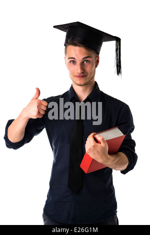 Heureux jeune homme diplômé de l'université, avec graduation hat et gros livre dans une main, faisant signe de pouce Banque D'Images