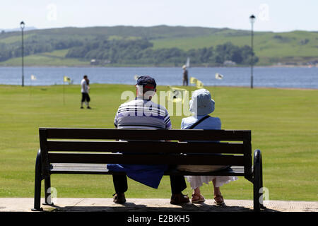 Largs, North Ayrshire, Écosse, Royaume-Uni, mercredi, 18 juin 2014. Lors d'un autre après-midi d'été sec et chaud, un couple s'assoit et se détend sur un banc de bord de mer tout en regardant les gens jouer un match de pitch and putt sur le Mackerston Putting Green dans la ville à côté du Firth of Clyde Banque D'Images