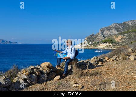 L'Italie, la Sicile, les îles Egadi, l'île de Levanzo, Cala Fredda, randonneur assis sur un rocher la lecture d'une carte sur un sentier de randonnée à l'île de Favignana dans l'arrière-plan Banque D'Images