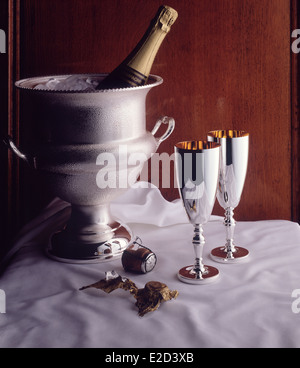 Une bouteille de champagne dans un seau à glace d'argent couverts de condensation,sur un linge blanc avec deux gobelets d'argent contre le bois. Banque D'Images