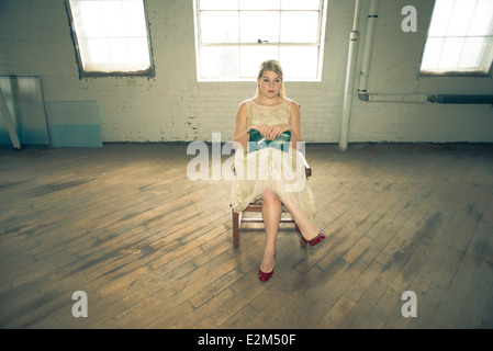 Un 16-year-old woman femme portant une robe en dentelle des années 60 tenant un sac à main vert vintage est assis sur une chaise dans un corset en usine. Banque D'Images