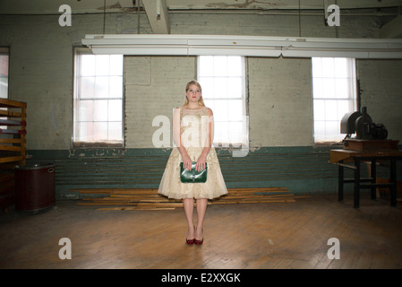 Un 16-year-old woman femme portant une robe en dentelle des années 60 tenant un sac à main vert vintage se trouve dans une chambre dans un corset en usine. Banque D'Images