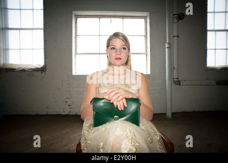 Un 16-year-old woman femme portant une robe en dentelle des années 60 tenant un sac à main vert vintage est assis sur une chaise dans un corset en usine. Banque D'Images