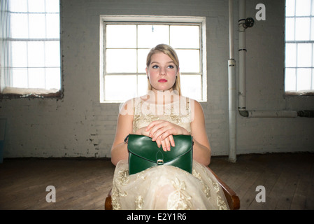 Un 16-year-old woman femme portant une robe en dentelle des années 60 tenant un sac à main vert vintage est assis sur une chaise dans un corset en usine. Banque D'Images