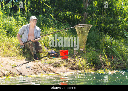 Personnes âgées handicapées pêcheur avec une jambe à l'aide d'un filet à la terre un poisson comme il est assis sur le sol au bord d'un lac Banque D'Images