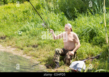 Homme handicapé avec sur jambe e assis sur la rive du lac casting sa canne à pêche qu'il bénéficie d'une journée de pêche dans le soleil de l'été Banque D'Images