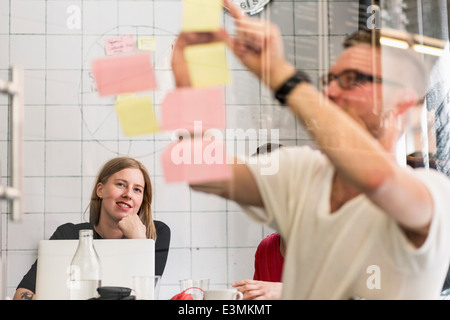 Jeune homme d'écrire leurs idées sur des notes adhésives avec des collègues en arrière-plan à creative office Banque D'Images