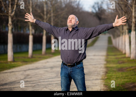 Homme d'âge moyen le soleil brille, debout avec ses bras étendu et les yeux fermés face au soleil dans un chemin rural bordé d'arbres Banque D'Images