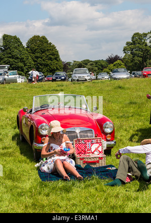 L'est du Devon, Angleterre. Une fete et Garden Party avec un couple assis au soleil à côté de leur voiture de sport MG rouge vintage. Banque D'Images