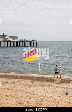 Southwold pier avec life guard drapeau patrouille de sécurité et 2 personnes sur la plage. Femme tenant une pelle rouge. Un château de sable bleu moule. Banque D'Images