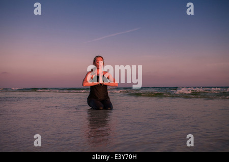 Femme mature pratiquer yoga pose agenouillée sur la plage au coucher du soleil Banque D'Images