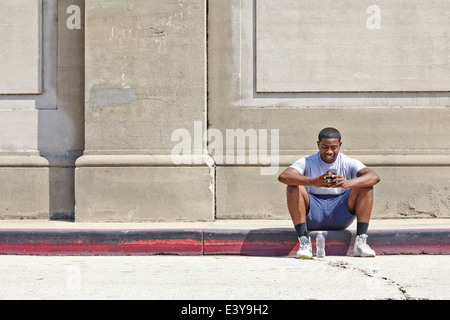 Young male runner assis sur trottoir texting on smartphone Banque D'Images