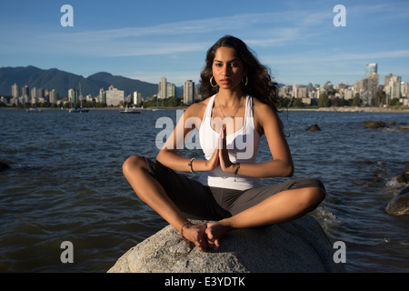 Une jeune femme indienne est de méditer sur un rocher avec l'océan et le centre-ville de Vancouver, à l'arrière-plan lointain. Banque D'Images