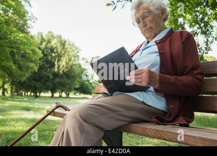 Senior woman sitting on park bench reading bible Banque D'Images