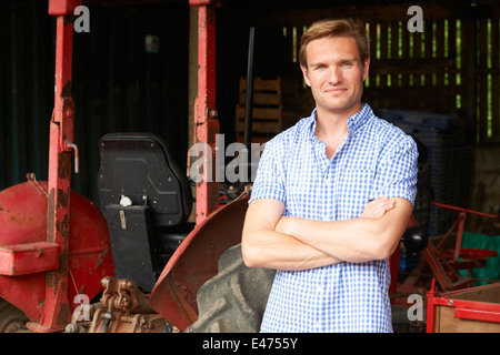 Farmer Standing Next to Old Fashioned Tracteur In Barn Banque D'Images