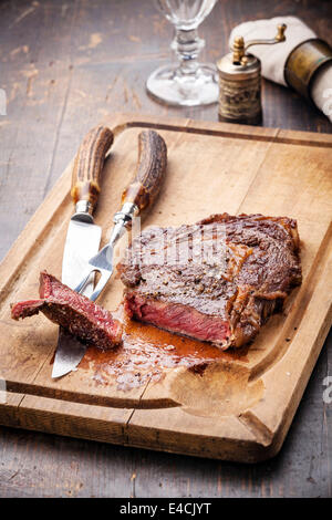 Pavé de boeuf grillée saignante Ribeye avec fourchette et couteau pour la viande sur une planche à découper sur fond de bois foncé Banque D'Images