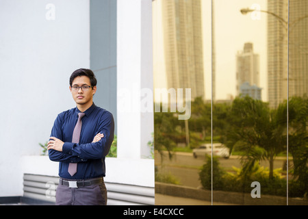 Portrait of Chinese businessman standing with arms crossed près de l'immeuble de bureaux à Panama. Réflexions de miroir sur la ville Banque D'Images